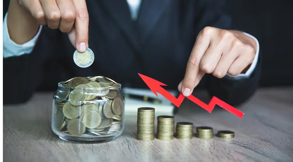 Mãos colocando moedas em um pote de vidro ao lado de pilhas de moedas e uma seta vermelha de crescimento