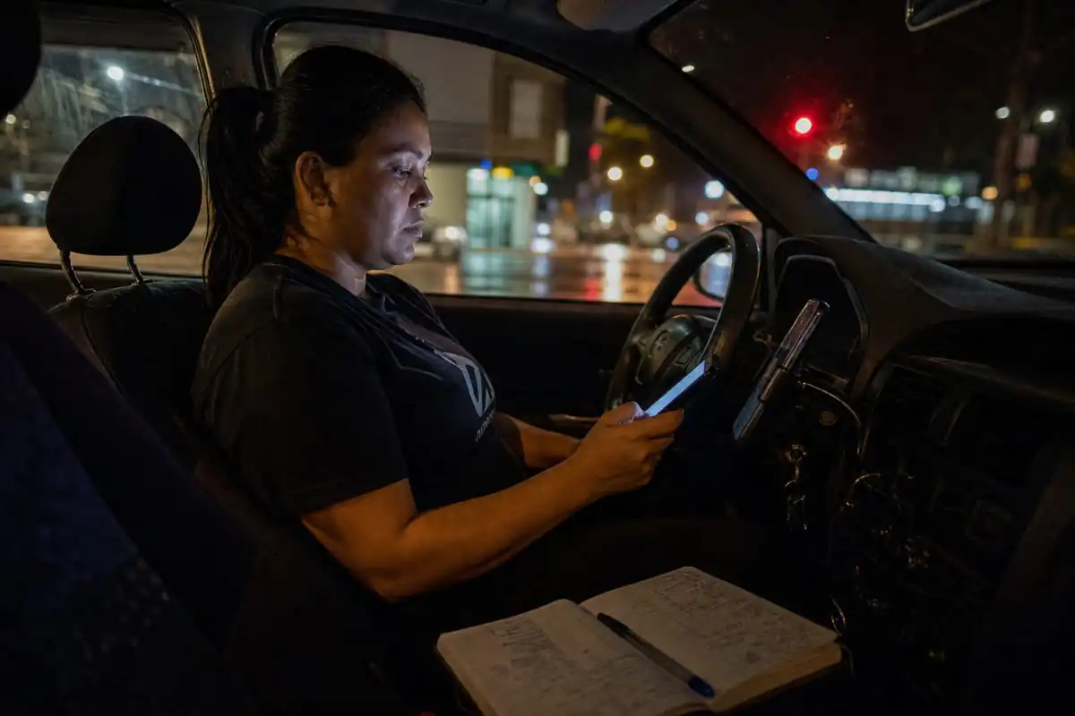 Motorista de aplicativo à noite dentro do carro, rosto iluminado pela luz do celular no suporte, com expressão de cansaço e indignação contida ao conferir o app do banco, caderno de anotações de gastos aberto no banco do carona