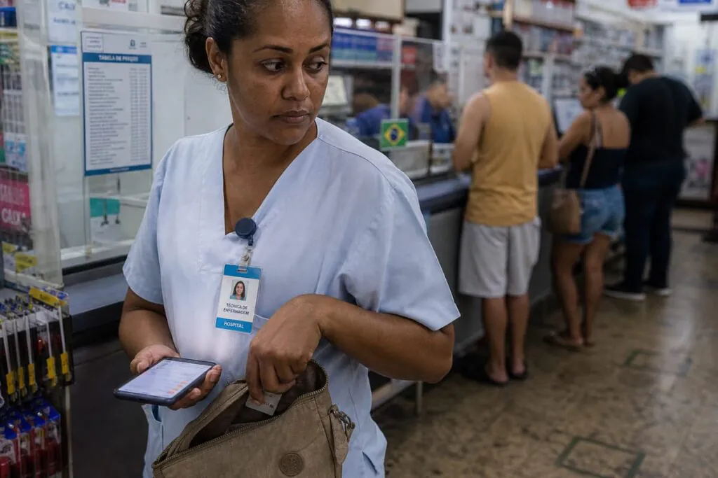 Técnica de enfermagem ainda de uniforme, com crachá do plantão, segura o celular baixo escondendo a tela com pendências negativadas enquanto guarda o cartão da mãe na bolsa, na fila de um correspondente bancário.